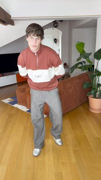 Brun og hvit zip-up jakke med logo, grå vid bukse og sølvfargede joggesko. Innendørs miljø med plante og teksturert sofa.