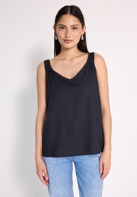 Młoda kobieta z długimi ciemnymi włosami, nosząca srebrne kolczyki w kształcie kół, czarną top bez rękawów z dekoltem w kształcie litery V oraz jasnoniebieskie dżinsy, stojąca na jednolitym tle.