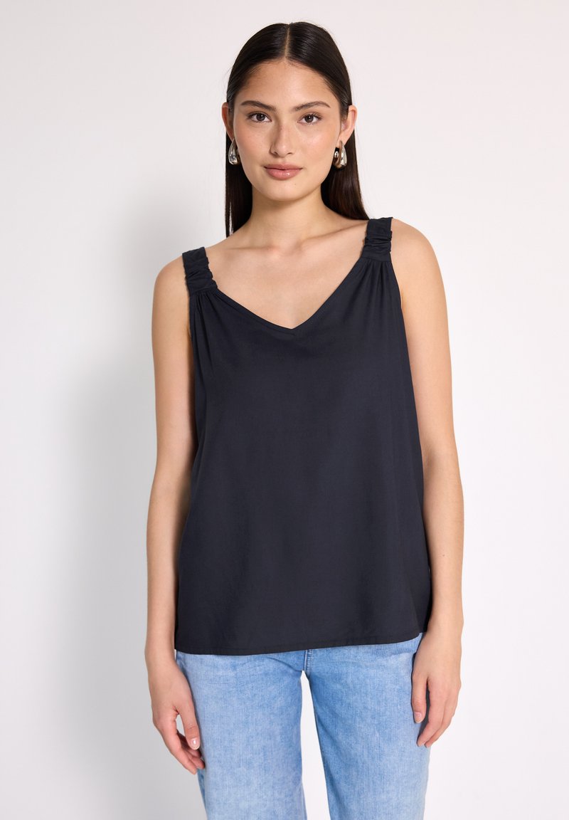 Młoda kobieta z długimi ciemnymi włosami, nosząca srebrne kolczyki w kształcie kół, czarną top bez rękawów z dekoltem w kształcie litery V oraz jasnoniebieskie dżinsy, stojąca na jednolitym tle.