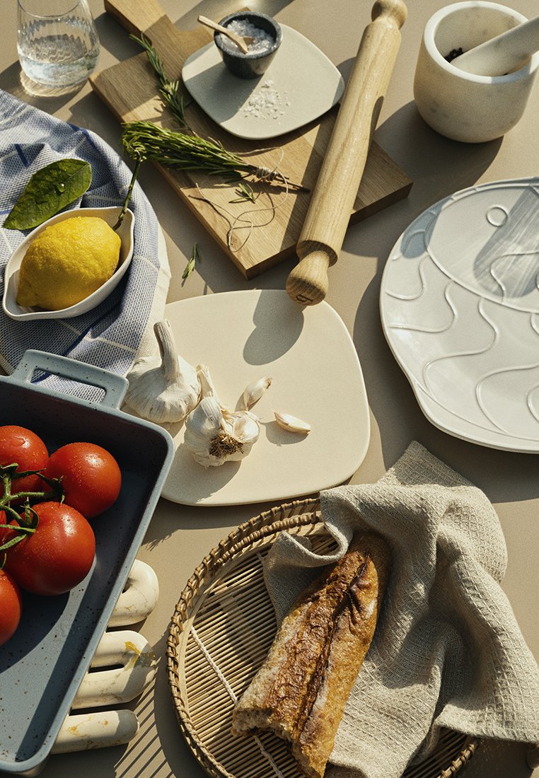 Een verscheidenheid aan keukenspullen gerangschikt op een oppervlak: tomaten, knoflook, een citroen, een deegroller, een gevlochten mand en een geweven linnen doek. Witte en grijze borden.