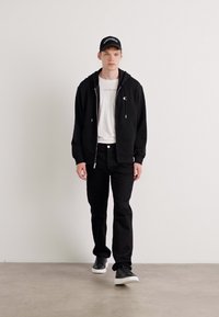 Sudadera negra con cremallera sobre una camiseta blanca, jeans negros y zapatillas negras. Los accesorios incluyen una gorra negra con texto. Texturas suaves en todo.