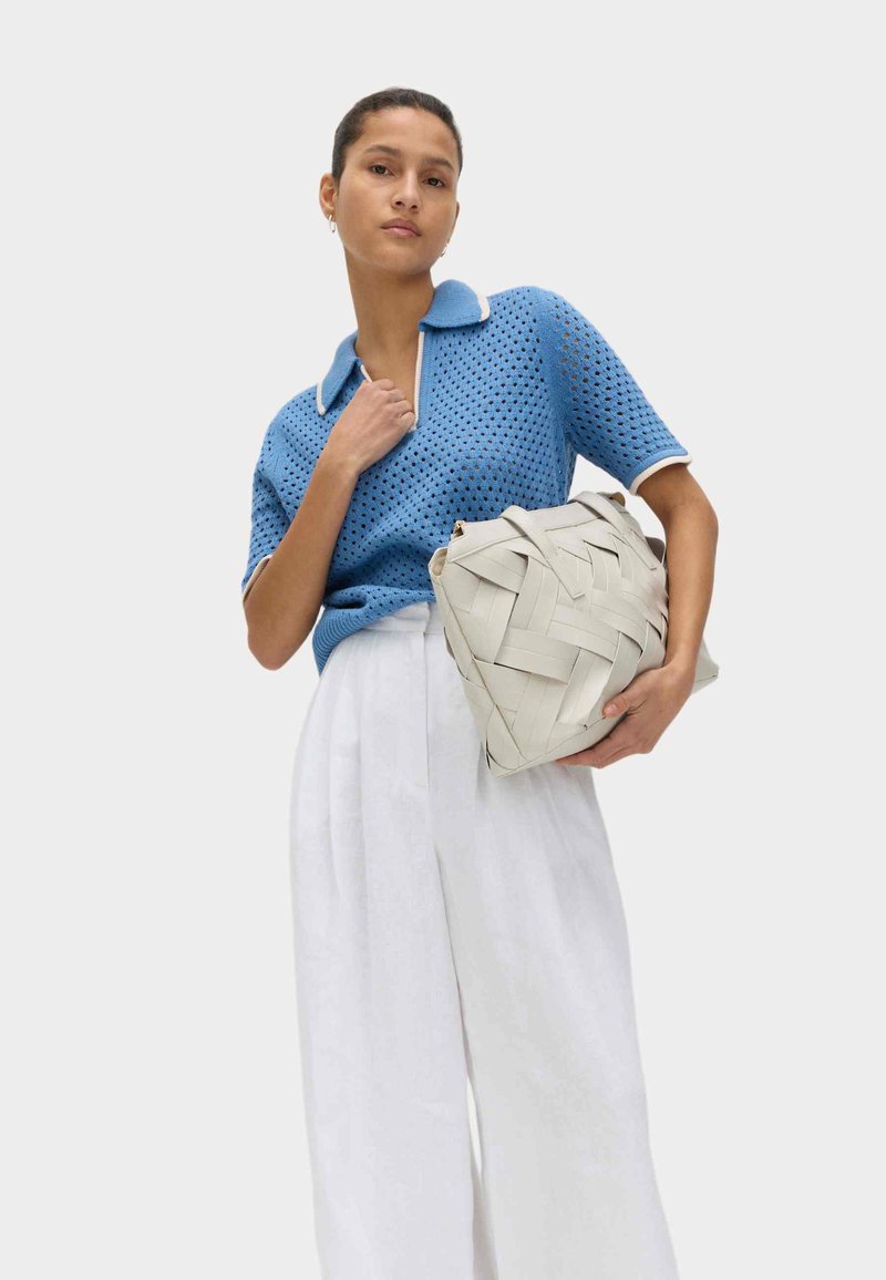 Femme portant un haut en maille bleu à manches courtes et un pantalon blanc à jambes larges, tenant un grand sac à main tissé beige sur un fond uni.