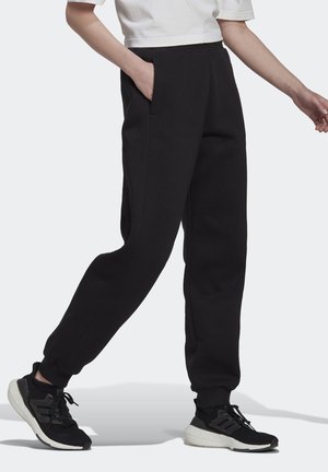Person trägt schwarze Jogginghosen und schwarze Laufschuhe mit weißen Sohlen, geht mit einer Hand in der Tasche und einem weißen, kurz geschnittenen Top.