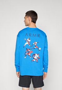Mėlyna ilgos rankovės marškinėlius su dinamiškų sportininkų grafika ant nugaros ir tekstu "ARMR" virš dizaino.