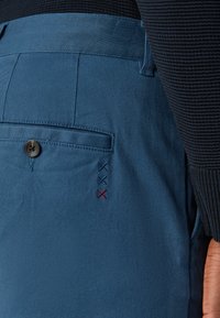 Kék nadrág hátulján egy gombos zsebből áll, és kék és piros dekoratív varrással rendelkezik. Az anyag sima, textúrált megjelenésű.