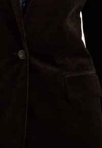 Ciemnobrązowa marynarka ze sztruksu z pionowymi prążkami, zapinana na jeden guzik, z klapkową kieszenią na dolnej przedniej części.