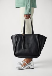 Borsa a tracolla in pelle nera texturizzata con un accento a frange, dotata di due manici robusti. Modello che indossa scarpe con tacco argento e pantaloni color crema.