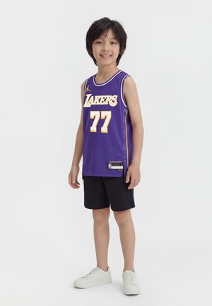 Egy fiú áll, aki lila Los Angeles Lakers kosárlabda mezt visel 77-es számmal, fekete rövidnadrágot és fehér tornacipőt, miközben mosolyog a kamerába.