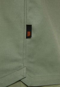 Groene stof met een gladde textuur, voorzien van een zwart label met een oranje vierkante accent nabij de zoom en schone stikdetails.