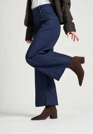 Pantalones de mezclilla de pierna ancha azul oscuro con costuras blancas, con una cintura alta y bolsillos, combinados con botas de ante marrón de tobillo.