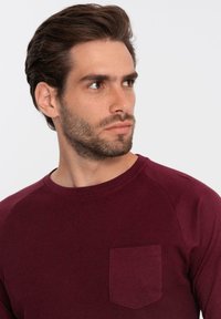 Camisa de manga larga burdeos con cuello redondo y un bolsillo en el pecho. La tela es texturizada y presenta acentos de hombros lisos en contraste.