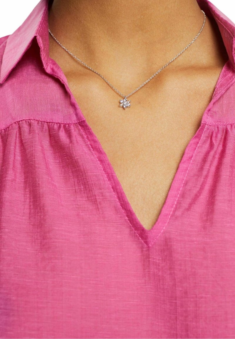 Collier en argent avec un petit pendentif floral porté autour du cou d'une personne, assorti à un chemisier rose à col en V.