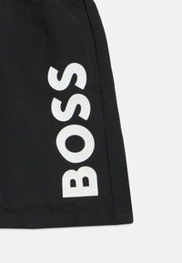 Juodi šortai iš minkštos medžiagos, kuriuose šone yra didelis baltas užrašas "BOSS". Yra šoninė kišenė.