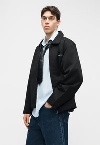 Νεαρός άνδρας που φορά μαύρο σακάκι, ανοιχτό μπλε πουκάμισο, φαρδιά μαύρη γραβάτα και σκούρο μπλε τζιν παντελόνι, στέκεται μπροστά σε λευκό φόντο.