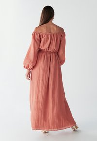 Ilga suknelė iš minkštos koralinės spalvos audinio, su nuogais pečiais, ilgomis pūstomis rankovėmis, elastinga liemeniu ir lengvai krentančia sijono dalimi iki čiurnos.