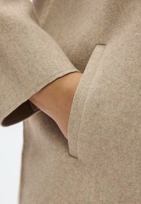 Μπεζ παλτό από τσόχα με απαλότητα στην υφή, με διπλωμένο μανσέτα και πλαϊνή τσέπη που καλύπτει εν μέρει το χέρι. Μινιμαλ ιστικός σχεδιασμός με καθαρές γραμμές.