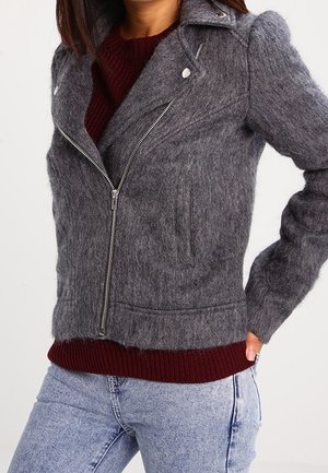 Femme portant une veste zippée grise ajustée et duveteuse par-dessus un pull bordeaux et un jean bleu clair, debout avec une main sur la hanche.