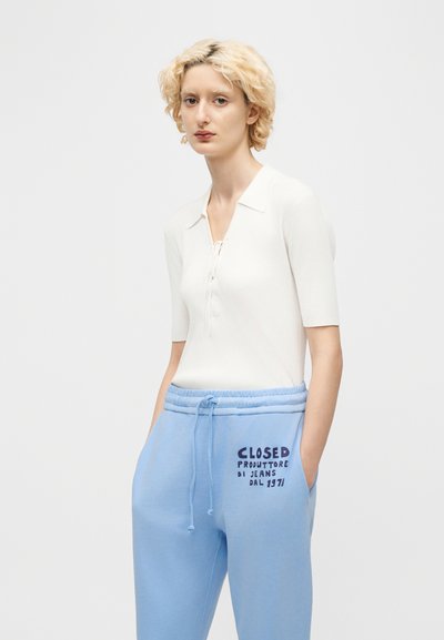 Άτομο με κοντά ξανθά μαλλιά που φοράει λευκό κοντομάνικο μπλουζάκι και ανοιχτόχρωμο μπλε φόρμες με κείμενο στο αριστερό μηρό.