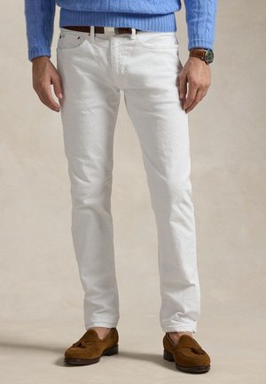 Homme portant un jean slim blanc, un pull en maille torsadée bleu, une ceinture en cuir marron, des mocassins en daim marron à pampilles, et une montre-bracelet au poignet gauche.