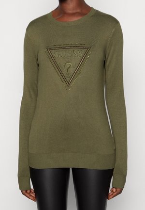 Kobieta nosi oliwkowozielony sweter z długim rękawem z teksturowanym logo Guess i trójkątnym zdobieniem w postaci ćwieków z przodu, zestawiony z czarnymi spodniami.