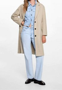 Béžový vlněný kabát přes světle modrou džínovou bundu a džínové kalhoty ve stejném odstínu. Černé boty doplňují outfit. Na bundě jsou viditelné knoflíky.