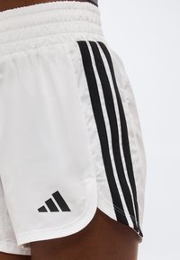 Λευκά αθλητικά σορτς με μαύρο σχέδιο με τρεις ρίγες στο πλάι και λογότυπο Adidas κοντά στο τελείωμα, που φοριούνται από άτομο με σκουρόχρωμο δέρμα.