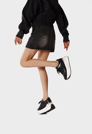 Donna che indossa una maglia nera a maniche lunghe, una minigonna di jeans scuro e sneakers nere con plateau, in piedi su una gamba con l'altra piegata all'indietro.