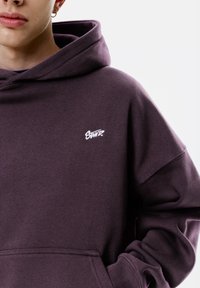 Sweat à capuche violet en tissu doux. Présente un petit logo brodé blanc sur la poitrine. Comprend une poche kangourou et des poignets côtelés.