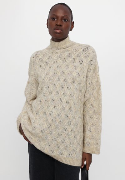 Beige gebreide coltrui met een gestructureerd ruitenpatroon, losse pasvorm en geribbelde cuffs. Gestyled met donkere broeken en vastgehouden door het model.