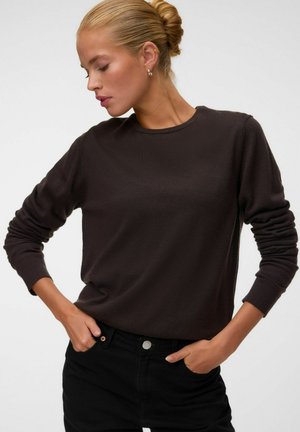 Donna bionda con capelli raccolti in una crocchia indossa un maglione marrone scuro a maniche lunghe e pantaloni neri con le mani nelle tasche, guardando in basso verso sinistra.
