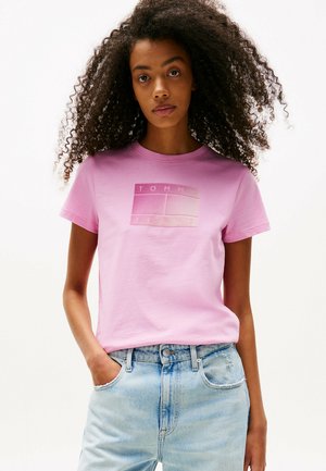 Camiseta de algodón rosa con mangas cortas que presenta un logo texturizado en el centro que dice "TOMMY JEANS." Combinada con jeans desgastados de color azul claro.