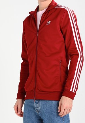 Persoon die een rode Adidas jas met ritssluiting draagt, met witte strepen op de mouwen en blauwe spijkerbroek, staand tegen een witte achtergrond.