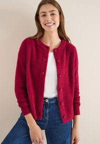 Karminroter strukturierten Cardigan mit Rundkragen, Knopfverschluss und gerippten Bündchen, kombiniert mit einem weißen Oberteil und blauen Jeans.