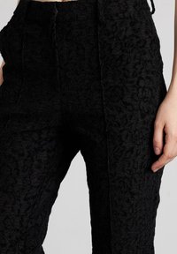 Pantalones negros hechos de una tela texturizada con un patrón floral, con una cintura alta y un ajuste relajado, detalles de costura visibles y bolsillos laterales.