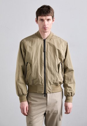Jachetă bomber ușoară, de culoare khaki, cu manșete și talie din țesătură ribbed. Prezintă o închidere cu fermoar și buzunare laterale cu deschideri în unghi.