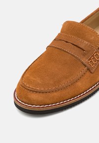 Bruine suède instappers met een gestikt ontwerp, voorzien van een gevoerd voetbed en een rubberen zool, geaccentueerd door een contrasterende witte naad.