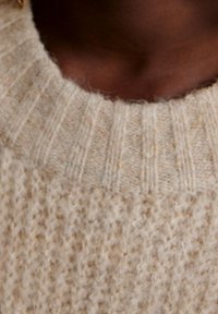 Maglione lavorato a maglia beige con un motivo testurizzato, colletto a coste e materiale morbido e peloso. Primo piano del collo e della zona della spalla.