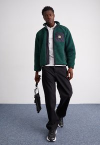 Grüne Fleece-Jacke mit einer schwarzen Reißverschlusstasche, darunter ein weißes Hemd, schwarze Hosen und graue Turnschuhe. Modell hält einen Regenschirm.