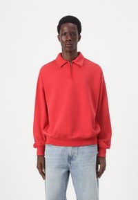 Pullover rosso in pile con colletto e zip a metà, con spalle scese e polsini a costine. Abbinato a jeans azzurri chiari.