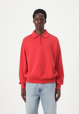 Red fleece pullover with a collar and half-zip, featuring drop shoulders and ribbed cuffs. Paired with light blue jeans.