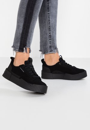 Person iført sorte plateau-sneakers med tykke såler og mørkegrå slidte jeans med frynset kant, stående på en hvid overflade.