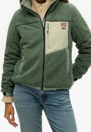 Person iført grøn fleecejakke med beige brystlomme og lyseblå jeans, stående med den ene hånd i lommen og den anden nedad.