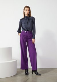 Blouse en soie bleu foncé avec un col cravate et des manches longues, associée à un pantalon large violet taille haute avec un détail de bouton. Bottes noires à talons aiguilles.