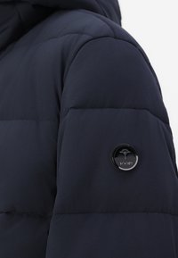 Jūras spēku pūkainā jaka uz plecu daļas ar quilted tekstūru, ar apaļu melnu pogu ar logotipu. Gluda auduma un pieguļošs siluets.