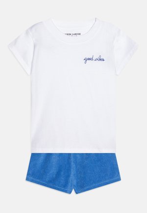 Hvid kortærmet t-shirt med "good vibes" tekst i blå på brystet kombineret med blå shorts på en ensfarvet baggrund.