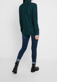 Grünes karriertes Hemd mit Button-Down-Kragen und Schulterklappen, kombiniert mit eng anliegenden dunkelblauen Jeans und schwarzen chunky Ankle Boots.
