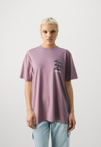 Camiseta de algodón morada con un corte relajado, que presenta diseños gráficos negros en el área del pecho izquierdo. Mangas cortas y cuello redondo.
