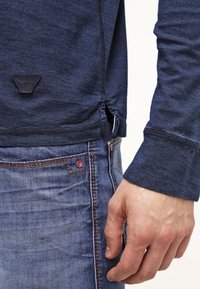 Námořnická košile s dlouhým rukávem, bočními rozparky, strukturovanou látkou a nášivkou s logem. V kombinaci s modrými džíny s kontrastním stehem a červeným akcentem.