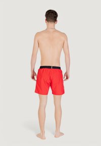 Homme debout pieds nus, de dos, portant un short de bain rouge vif avec une ceinture noire, sur un fond neutre.