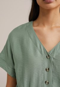 Top verde chiaro a maniche corte con scollatura a V, caratterizzato da quattro bottoni frontali e un tessuto testurizzato. Design pulito e semplice.